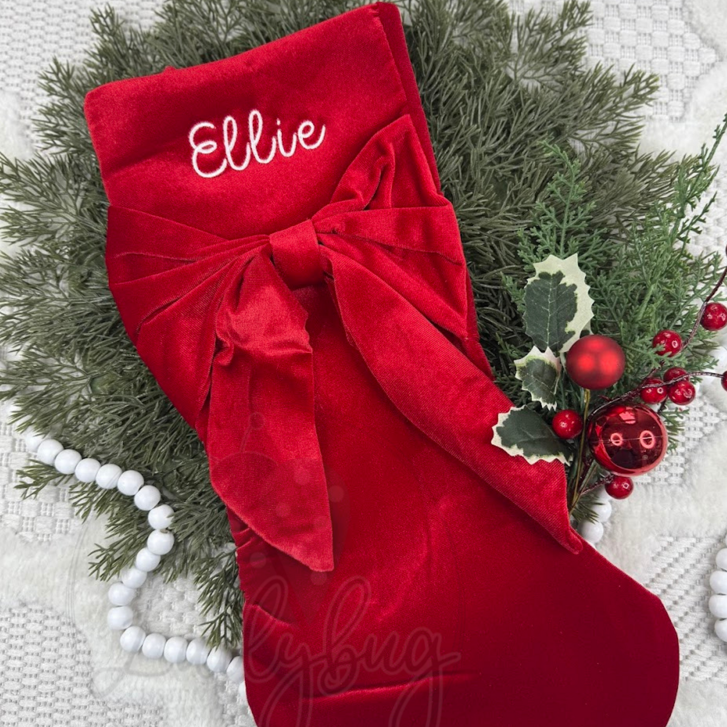 Red Christmas stocking with a large bow and personalized name, surrounded by greenery and berries.