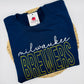 A folded navy blue crewneck t-shirt with 'milwaukee BREWERS' text across the front, placed on a round woven straw mat against a white background. The shirt features a white script 'milwaukee' and large outlined 'BREWERS' lettering in yellow.