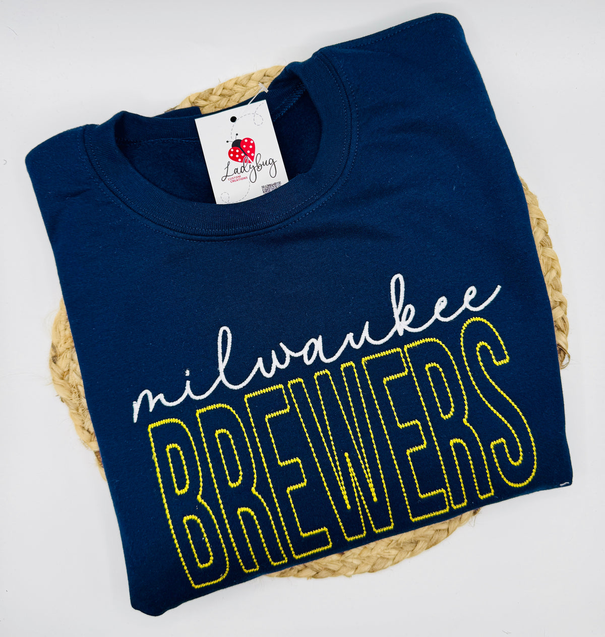 A folded navy blue crewneck t-shirt with 'milwaukee BREWERS' text across the front, placed on a round woven straw mat against a white background. The shirt features a white script 'milwaukee' and large outlined 'BREWERS' lettering in yellow.